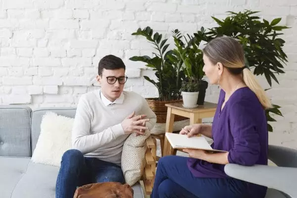 man--talking-about-his-problems-during-psychological-therapy-session-sitting-coach-while-mature-female-psychologist-with-copybook-listening-him-making-notes